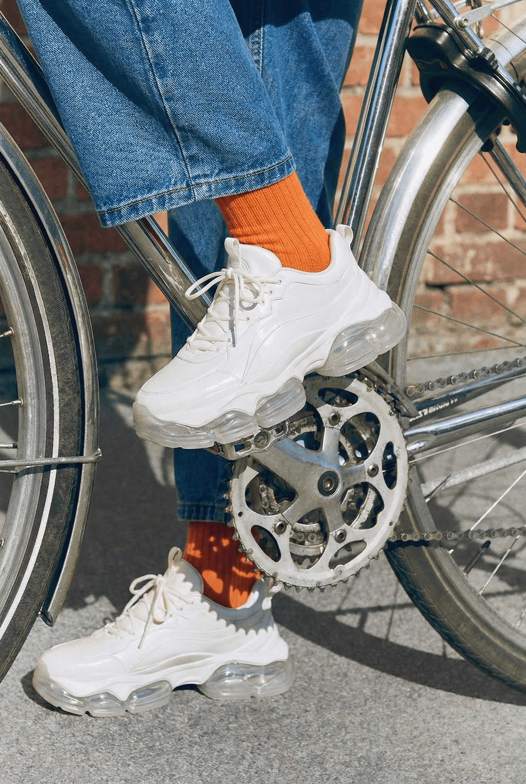 White chunky sneakers with orange socks and denim jeans styled for casual streetwear fashion beside a bicycle.