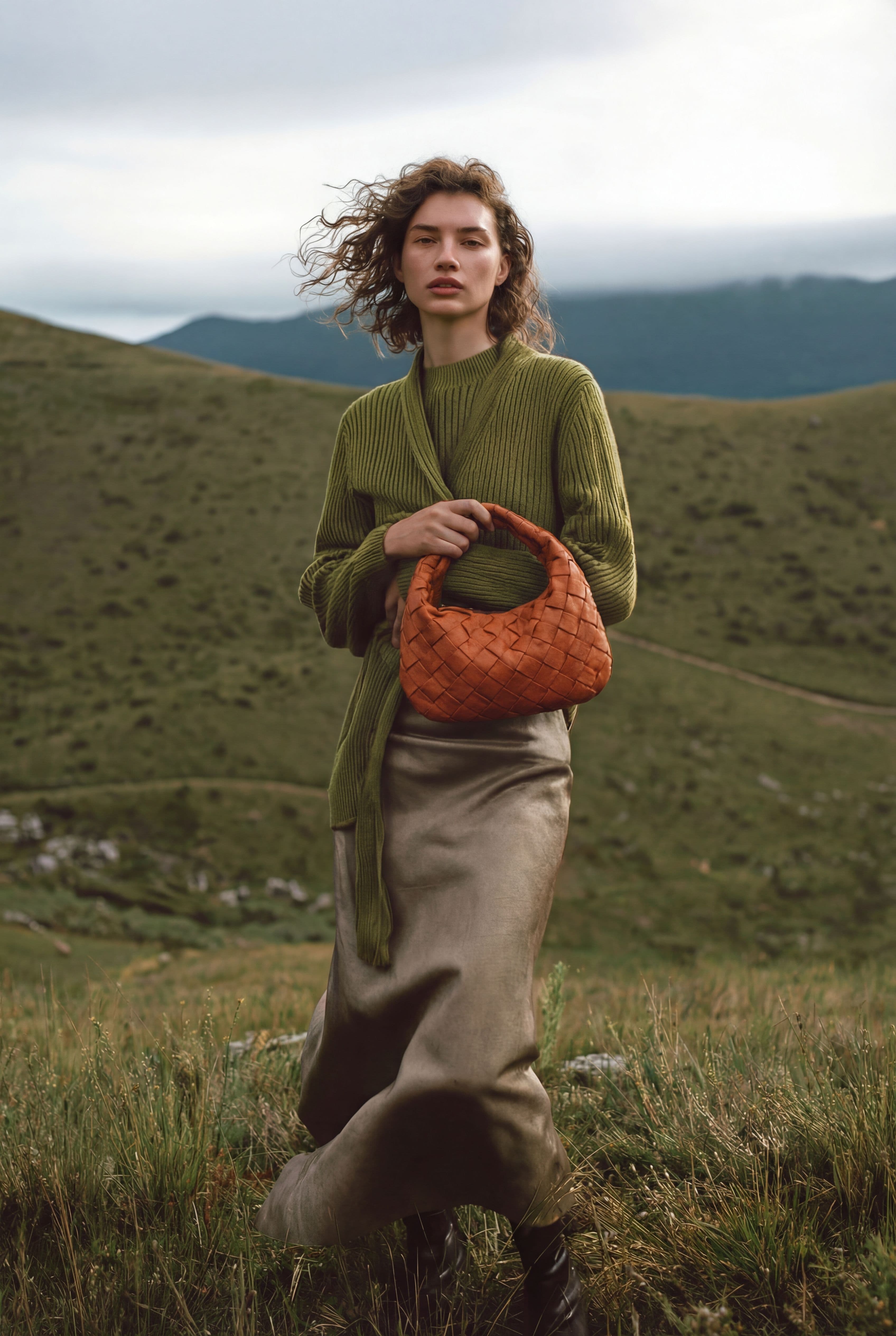 Woman holding a woven leather handbag with an olive knit sweater and satin skirt in a luxury fashion campaign outdoors.